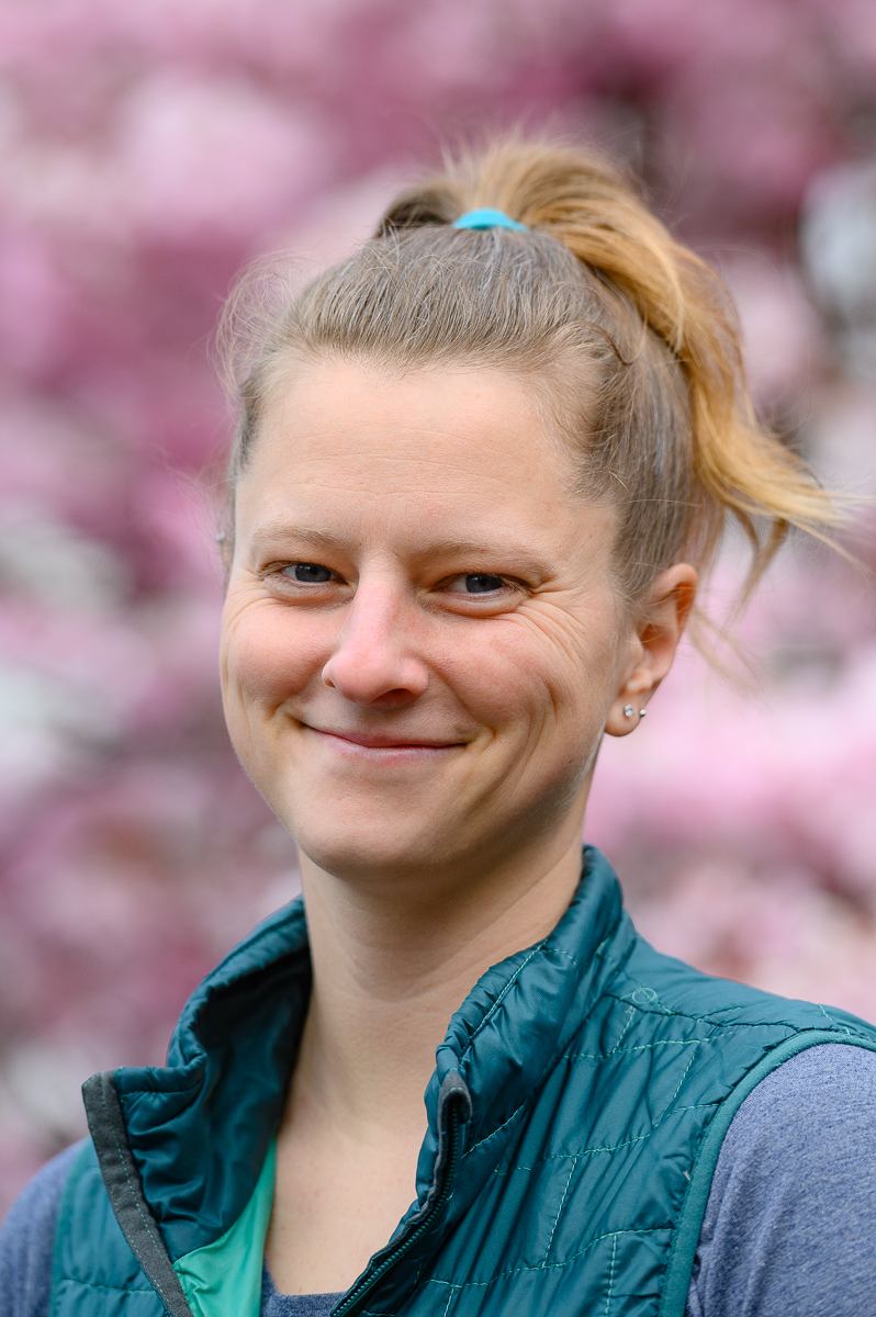 Bert, a white femme presenting person, stands in front of a blooming cherry tree.